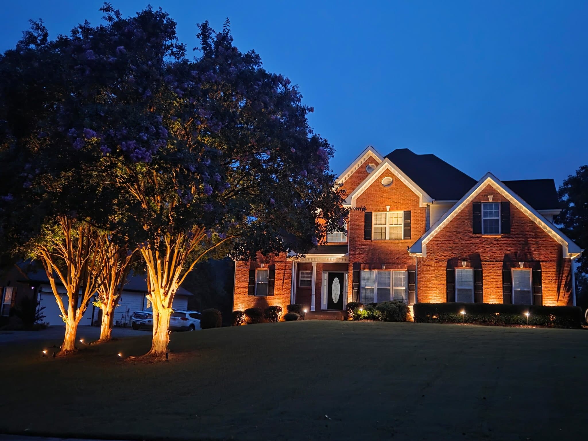 Two-story brick home in Hoschton, GA at blue hour with crape myrtle trees uplit in the front lawn, warm soffit lighting tracing the gables, and facade uplighting