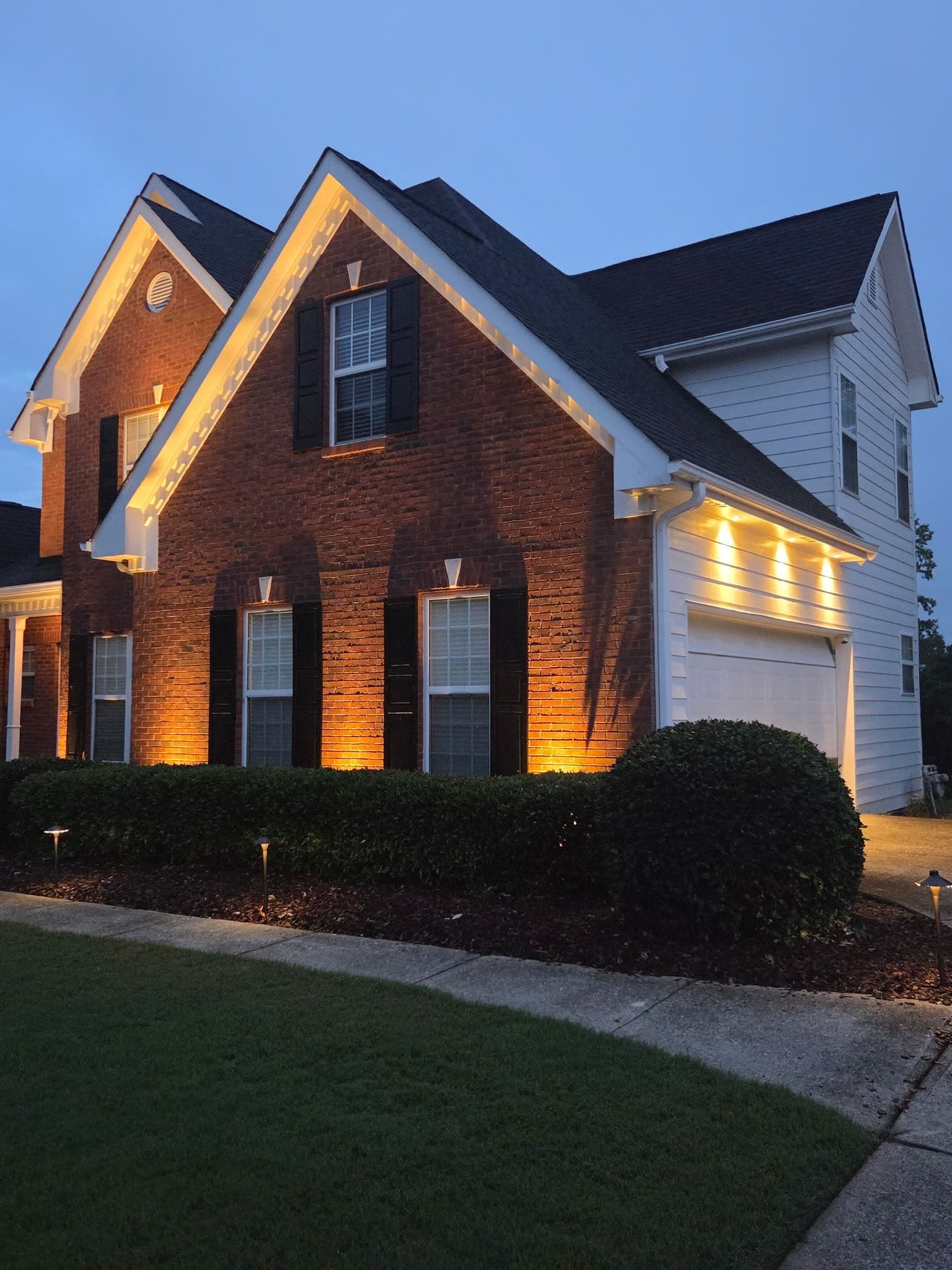 Two-story brick home in Hoschton, GA at dusk with warm soffit and eave lighting outlining the gable rooflines, downlights over the garage, and uplighting on the brick facade