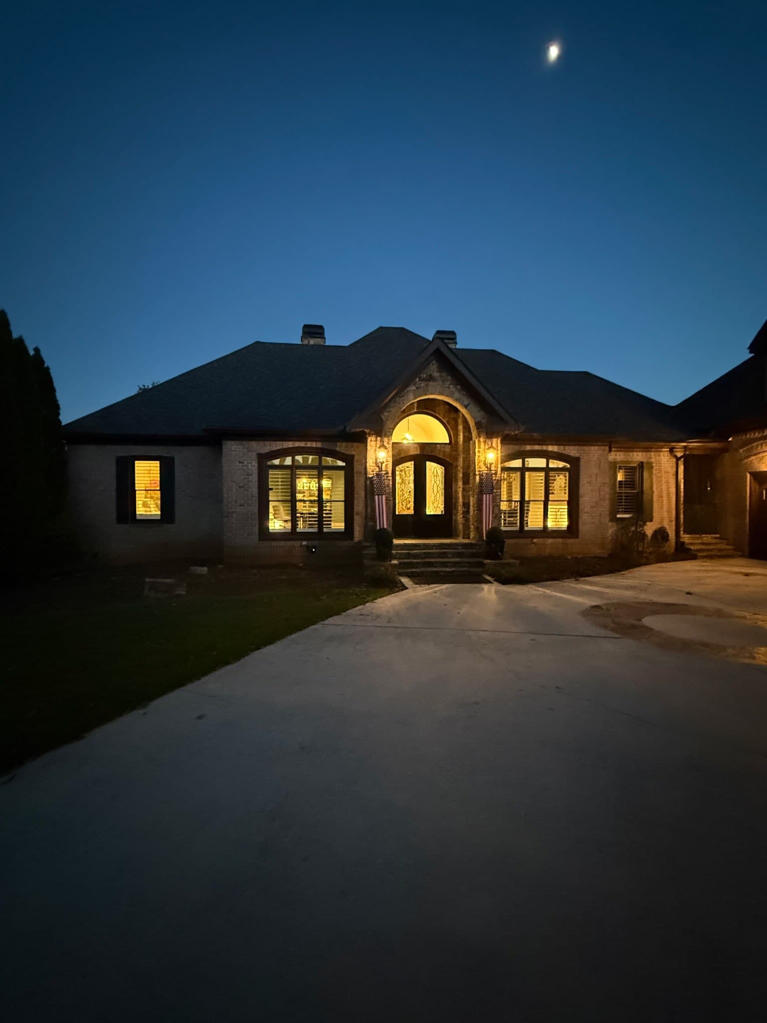 Brick home at night under a moonlit blue sky with a warmly lit arched stone entryway flanked by American flags