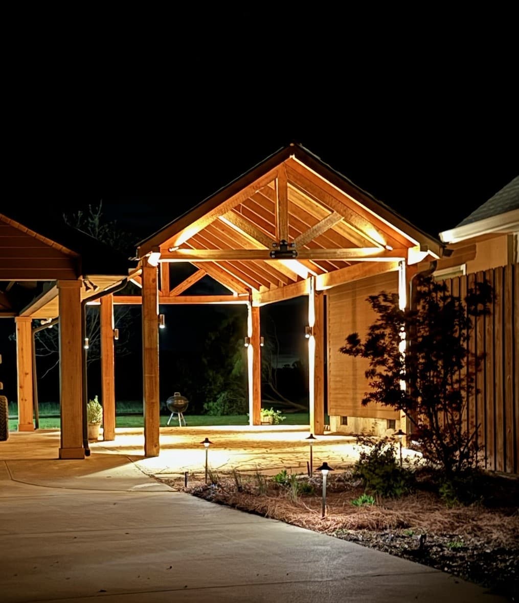Timber-frame pavilion lit along the rafters and posts over a paver patio at night