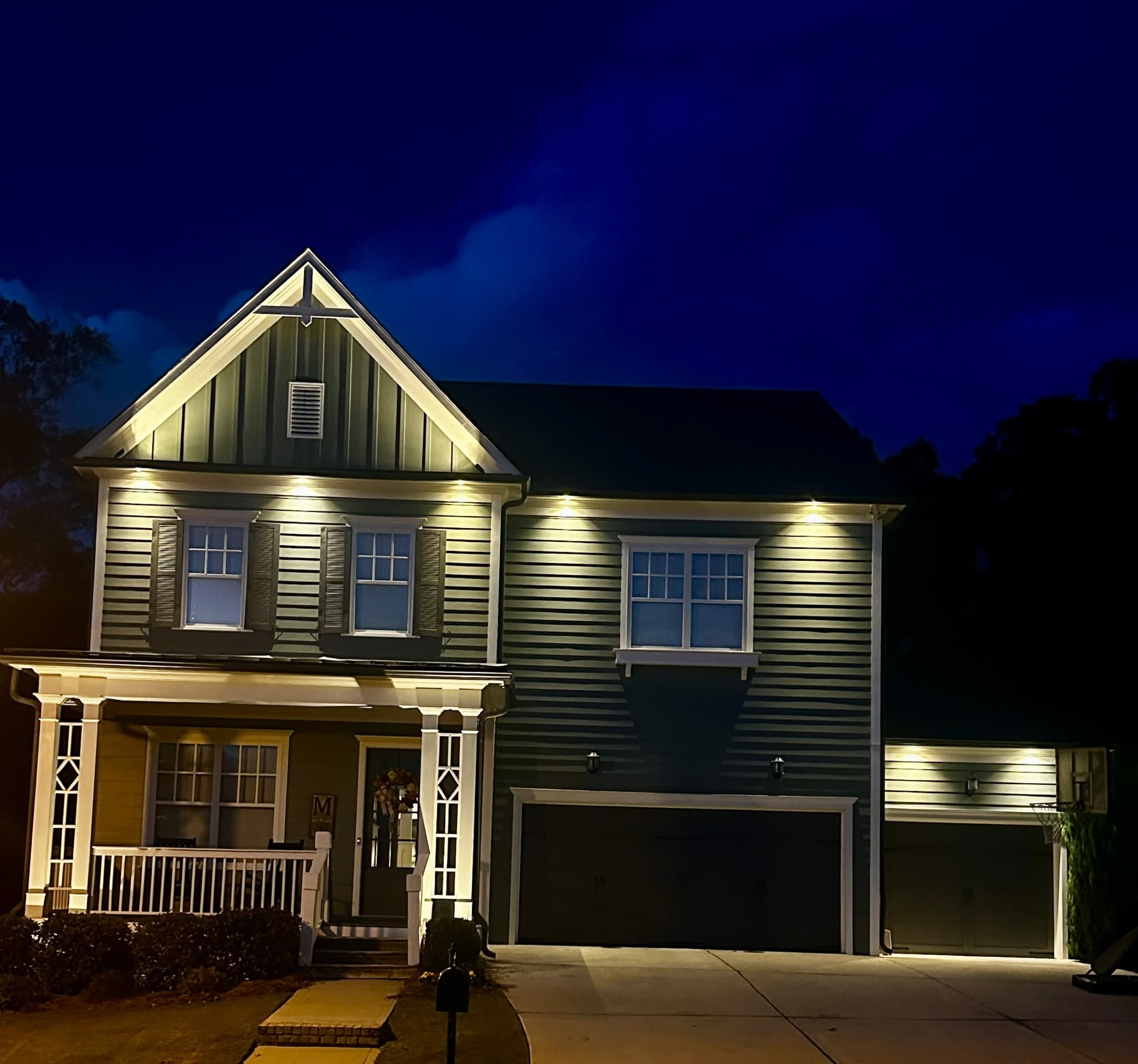 Two-story farmhouse at twilight with warm soffit downlighting washing the siding under the eaves and accenting the gable peak