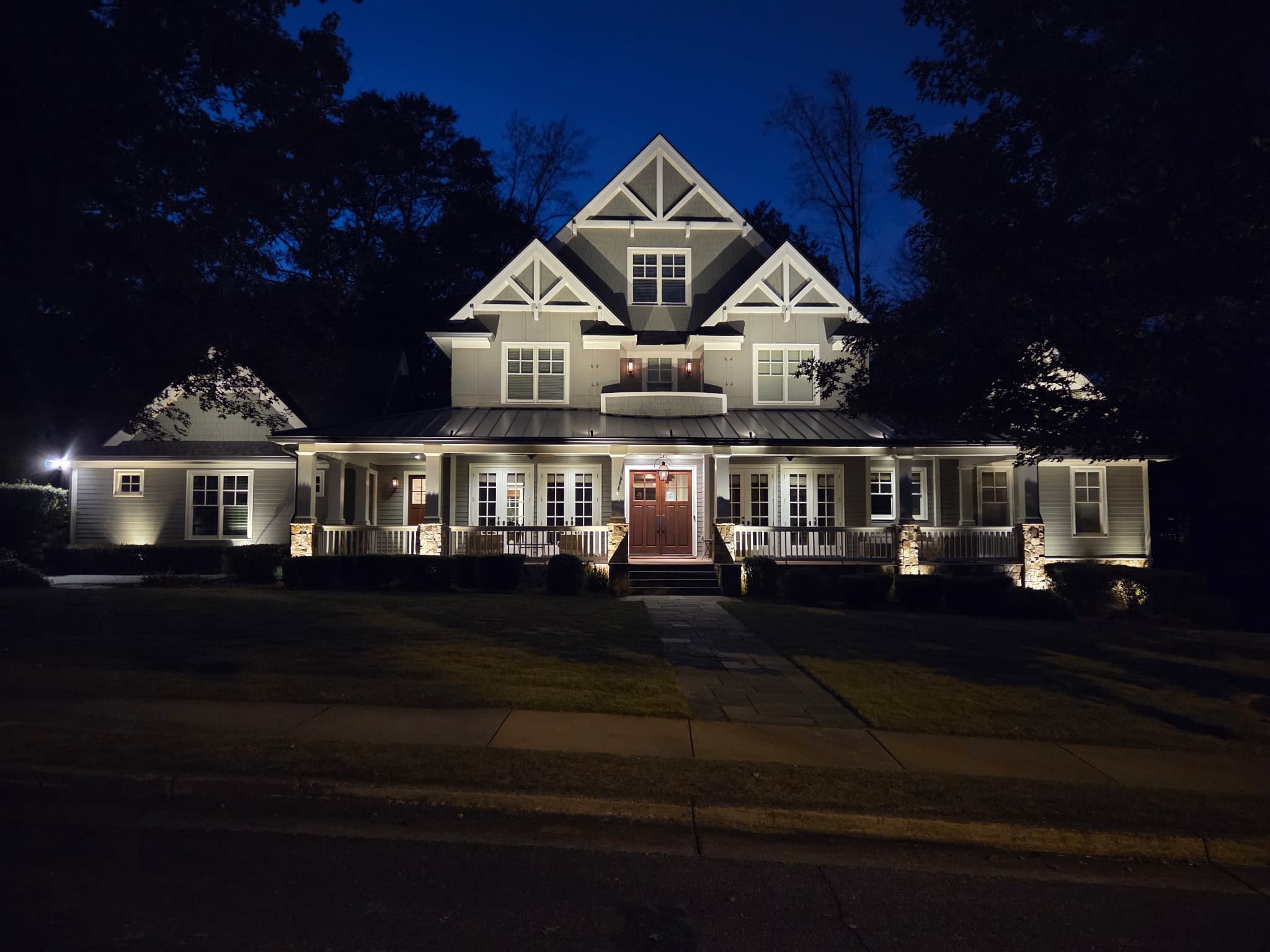 Grand two-story craftsman estate at night with dramatic uplighting on the gables, white timber trusses, and a stone wrap-around porch