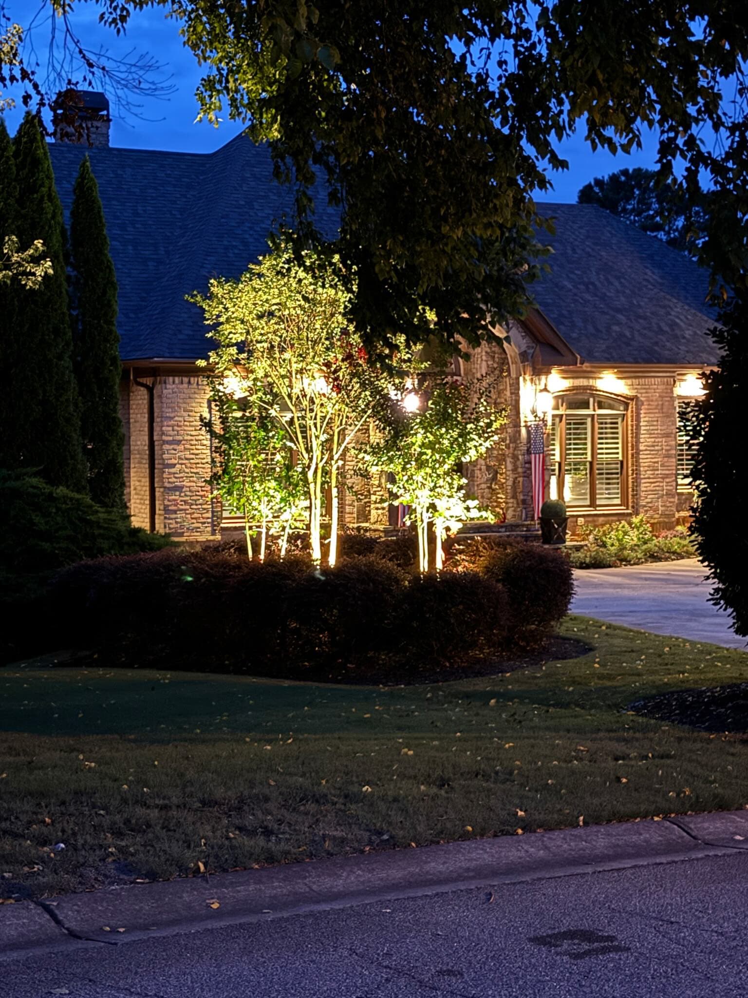 Stone and brick home at twilight with ornamental trees brightly uplit in the front bed and warm eave lighting across the facade