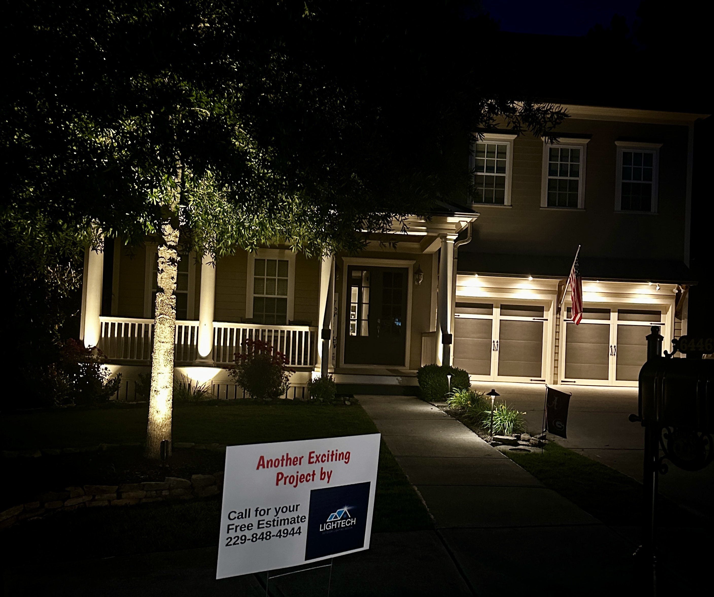 Two-story home at night with warm uplighting and a Lightech project yard sign in the foreground
