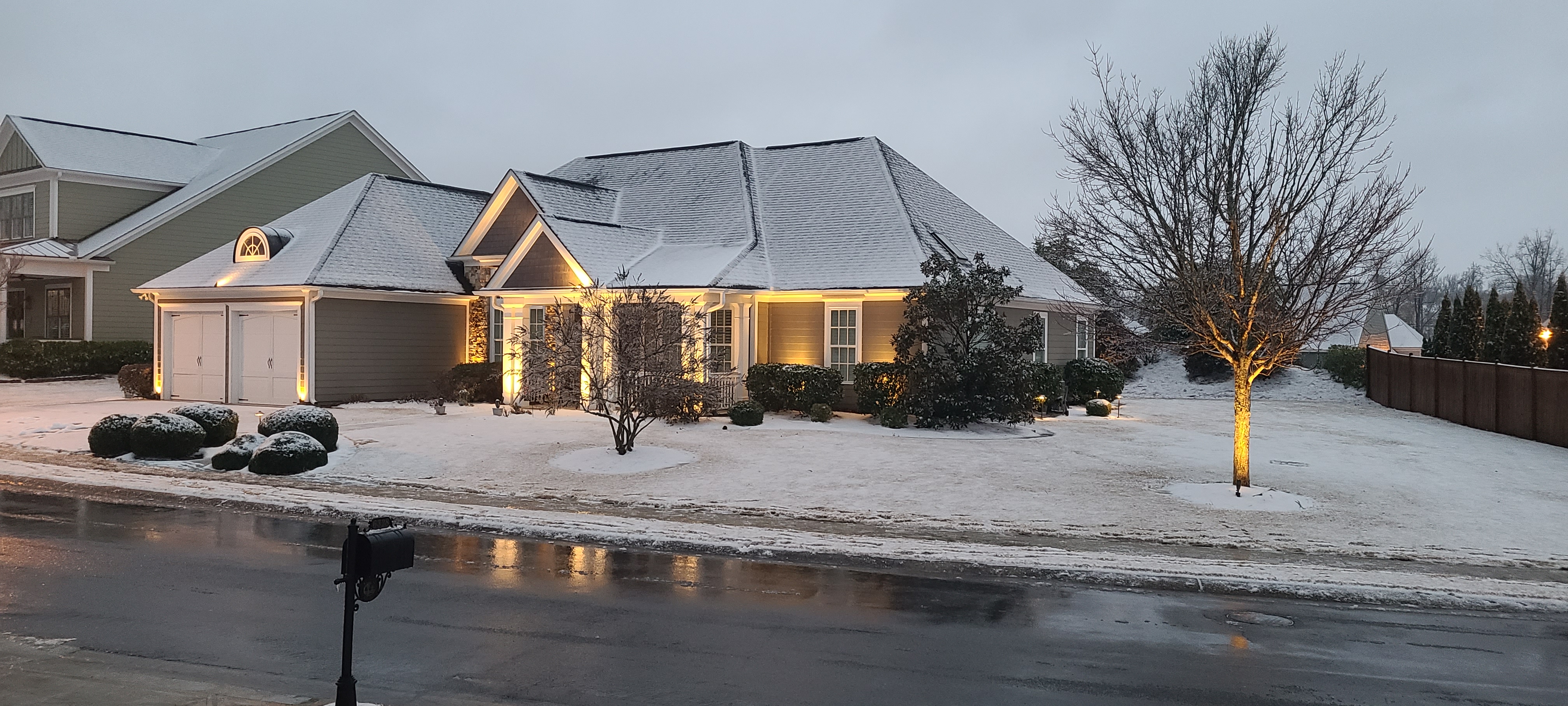 Green and tan ranch home in light snow with warm facade uplighting and an uplit bare tree
